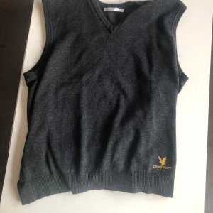 Mörkgrå stickad väst från Lyle & Scott - Snygg mörkgrå stickad väst från Lyle & Scott med gul broderad logga på nedre kanten. Västen har v-ringad hals och ribbade kanter för en clean look. Perfekt att slänga över en skjorta för extra stilpoäng.