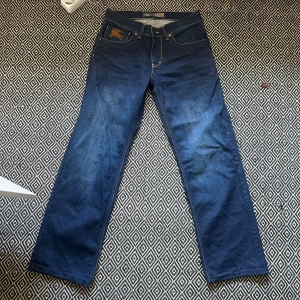 Blå jeans från Burberry - Snygga blå jeans från Burberry med klassisk rakt snitt och straight passform. Jeansen har bruna broderade detaljer och Burberry-logga på bakfickan. Materialet är slitstarkt jeans med kontrastsömmar. Superbra skick. W30 L34.