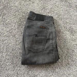 Grå jeans från Nudie Jeans Co - Säljer ett par grå jeans från Nudie Jeans Co med klassisk femficksdesign och svarta detaljer på bakfickan. Jeansen har rak passform och råa, fransiga benslut för en cool och avslappnad vibe. Materialet är mjukt denim i bomull med dragkedja framtill.