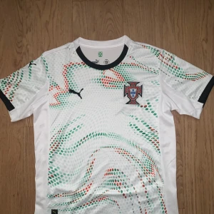 Portugal Puma vit fotbollströja S - Snygg Portugal fotbollströja från Puma i vit med prickigt mönster i grönt och rött. Kortärmad med svart krage och detaljer. Broderat Portugal-emblem på bröstet. Tillverkad i lätt och ventilerande polyester, perfekt för match eller träning.