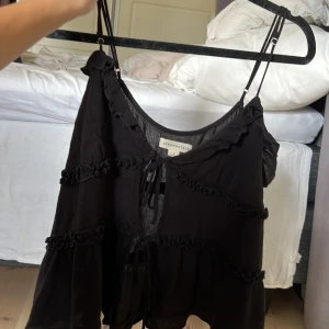 Svart linnetopp från Aeropostale - Svart linnetopp med tunna justerbara axelband och volangdetaljer från Aeropostale. Toppen har knytning framtill och flera dekorativa rynkade band som ger en cool och bohemisk vibe. Materialet är lätt och luftigt