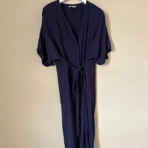 Marinblå långklänning från H&M - Snygg marinblå långklänning från H&M i storlek 42. Klänningen har v-ringad omlottfront, korta ärmar och ett knytband i midjan som ger en chill och avslappnad siluett. 