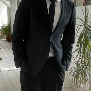 Svart kostym från Riley - Stilren svart kostym  med klassisk kavaj och kostymbyxor från Riley. Kavajen har två knappar, smal passform och diskreta fickor. Perfekt för dig som vill ha en clean och snygg look med tidlös vibe. Skjorta medföljer ej. Passar bra för dig som är mellan 180 till 190cm.