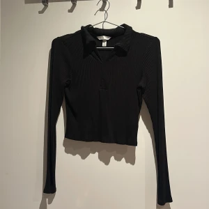 Svart ribbad croppad polotopp - Svart croppad polotopp med långa ärmar och krage. Toppen är ribbad och har en snygg v-ringning framtill. Perfekt för dig som gillar stilrena och trendiga plagg med lite edge.