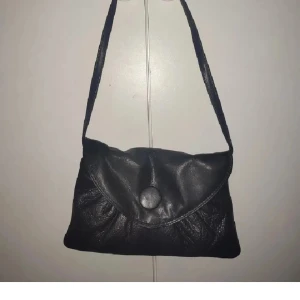 Svart axelväska i skinn deposhe - Snygg svart axelväska i skinn med enkel design och lock med stor knapp framtill. Perfekt storlek för det viktigaste och lätt att bära över axeln. Stilren och passar till många olika outfits.