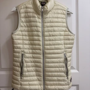 Colmar quiltad dunväst i ljus beige - Quiltad dunväst från Colmar i ljus beige med horisontella sömmar och hög krage. Västen har två fickor med dragkedja och en snygg Colmar-logga framtill. Perfekt att slänga över en hoodie för extra värme. Stängs med dragkedja framtill. Aldrig använd tyvärr pga för liten storken 🧸står ingen storlek men skulle kanske säga att den nassar xxs/xs
