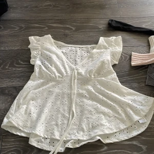 Vit blus med spets och volang XS - Supersöt vit blus från Zara i storlek XS med broderade spetsdetaljer och små hålmönster. Blusen har v-ringning, volangärmar och knyt framtill som ger en romantisk vibe. Perfekt för dig som gillar ljusa och luftiga toppar med extra detaljer.