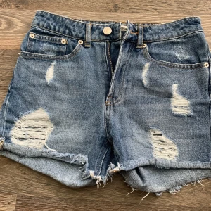Blå jeansshorts med slitningar Bik Bok - Snygga blå jeansshorts från Bik Bok i storlek XS. Modellen har hög midja, råa kanter och flera slitningar framtill och baktill för en cool, avslappnad vibe. Klassisk femficksdesign och knapp- samt dragkedja framtill. 