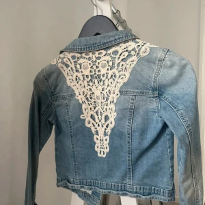 Jeansjacka med spets från Lab Industries - Snygg ljusblå jeansjacka från Lab Industries med unika spetsdetaljer i vitt på ryggen. Klassisk krage, bröstfickor med knappar och långa ärmar. Jackan har en croppad passform och knäppning framtill. Perfekt för dig som vill sticka ut med coola detaljer.