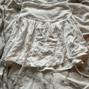 Vit kortkjol med volang från SHEIN - Supersöt vit kortkjol från SHEIN med volangkant och bred linning. Kjolen är gjord i ett lätt och luftigt bomullsmaterial, perfekt för varma dagar. Den har en snygg rynkad detalj bak och knytband i sidorna för extra stilpoäng. Tyvärr har tyget spruckit men annars är den i bra skick