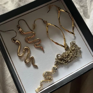 Guldiga örhängen och halsband med orm - Säljer ett set med två par guldiga örhängen, ett par i ormform och ett par med twistad, oval design. Medföljer även ett halsband med en glittrig ormberlock dekorerad med små stenar. Halsbandet har tappat lite av den guldiga färgen, men inte något som syns mycket 🥰finns endast baksidorna till ett par av örhängena. Säljer då de inte kommer till användning