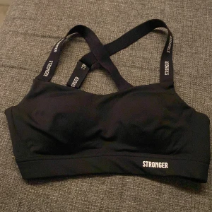 Svart sporttopp från Stronger - Supersnygg svart sport Bh från Stronger med korsade axelband där loggan är tryckt i vitt. Toppen är tight och har en sportig vibe, perfekt för gymmet eller andra träningspass. Materialet är mjukt och stretchigt💕💕💕💕Endast provad några gånger. Aldrig använt vid träning eller nånting💕💕💕ordinarie pris 599