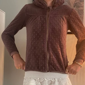 Lila fleecejacka med huva - Mysig brun fleecejacka med huva och dragkedja framtill. Jackan har ett prickigt mönster i materialet och ribbade muddar vid ärmsluten. Perfekt för kyliga dagar när du vill vara både bekväm och snygg.
