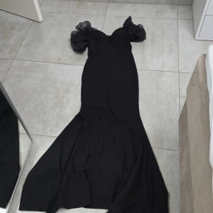 Svart långklänning med puffärmar - Säljer en elegant svart långklänning med dramatisk siluett och stora puffärmar i transparent tyg. Klänningen har en figurnära passform upptill och breder ut sig nedtill för en snygg look. Perfekt för dig som vill sticka ut med stilrena detaljer och volym vid axlarna.