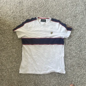 Vit t-shirt Lyle & Scott - Vit t-shirt från Lyle & Scott i storlek barn XL med mörkblå bred rand över bröstet och axlarna, samt röda detaljer längs ränderna. Klassisk rund hals och korta ärmar. Lyle & Scott-logga broderad på vänster bröst. Skön bomullskvalitet, perfekt för chill dagar.