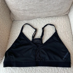 Svart sport-bh från Newbody - Svart sport-bh från Newbody med smala justerbara axelband och v-ringad front. Toppen är tillverkad i mjuk och stretchig polyesterblandning som sitter skönt mot kroppen. Perfekt för träning eller chill dagar när du vill ha något enkelt och bekvämt.