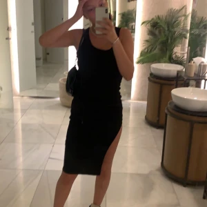 Svart ärmlös klänning med slits - Säljer en svart, ärmlös klänning med rund hals och en hög slits på sidan. Klänningen har en tight passform och är knälång, perfekt för dig som gillar en enkel men ändå edgy stil. Materialet känns mjukt och stretchigt mot kroppen.