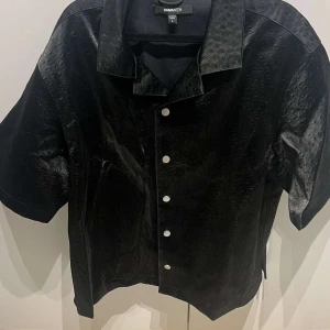 Svart skjorta från NovaMen med kort ärm - Svart skjorta från NovaMen i storlek L med korta ärmar och klassisk krage. Skjortan har en blank yta med subtilt prickigt mönster och vita knappar framtill. Passformen är loose och materialet har en syntetisk känsla som ger en cool look.