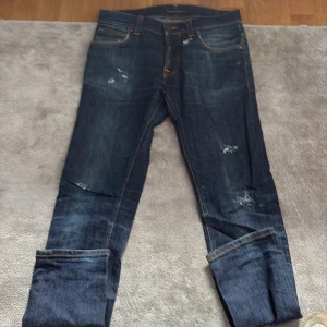 Mörkblå jeans från Nudie Jeans - Snygga mörkblå jeans från Nudie Jeans med klassisk femficksdesign och orangea sömmar. Jeansen har slitningar och en cool tvättad look, samt Nudie-logga på bakfickan. Passformen är rak och materialet är robust denim i bomull.