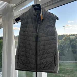 Grå quiltad dunväst från Parajumpers - Snygg grå dunväst från Parajumpers med quiltad design och hög krage. Västen har dragkedja framtill, sidofickor och en bröstficka med zip. Märkets patch syns på ryggen och insidan har en cool orange detalj. Perfekt för lager-på-lager och chill stil. Pris kan diskuteras vi en snabb affär.