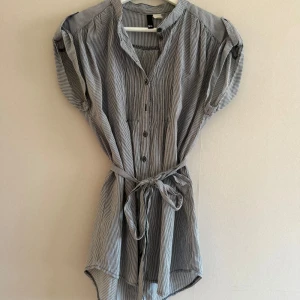 Randig blus med knyt i midjan - Snygg blus från H&M i svart och vit randigt mönster. Klänningen har knappar framtill, korta ärmar med knappdetalj och ett knytband i midjan för en chill passform. Fint skick och rökfritt hem!