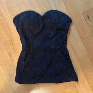 Svart spetsig korsett/ tub -topp - Snygg svart korsett-topp med hjärtformad urringning och spetsdetaljer. Toppen är axelbandslös och sitter tight mot kroppen för en ”hourglass look”. Köpt utomlands, unik! 