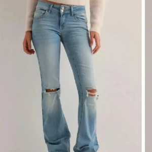 Bootcut ljusblå jeans med slitningar - Säljer ett par ljusblå bootcut jeans med snygga slitningar vid knäna och fransig nederkant. Modellen har klassiska fem fickor, inklusive bakfickor med lock och knapp. Jeansen har normal passform och är tillverkade i mjukt denimtyg som ger en skön känsla. Jeansen är nedsydda och har ett litet håll i slutet som inte syns när man har på sig dom. Skriv för fler bilder, och köp ej direkt. Storlek 36