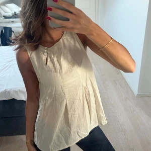 Krämvit ärmlös blus med knappar - Säljer en krämvit blus i luftig bomull med ärmlös design och två dekorativa knappar vid halsen. Blusen har lätt A-linjeformad siluett och ett litet nyckelhål framtill. Perfekt för varma dagar och enkel att styla med jeans eller shorts.