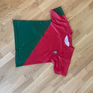 Portugal fotbollströja röd/grön - Säljer en Portugal fotbollströja med diagonal design i rött och grönt. Tröjan har korta ärmar och är gjord i ett lätt, syntetiskt material som andas. På bröstet finns Portugals landslagsemblem och på ryggen ett litet gult märke. Perfekt för match eller häng med kompisar.