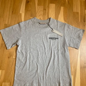 Grå Essentials Fear of God t-shirt - Snygg grå t-shirt från Essentials Fear of God med svart logotryck både fram och bak. Klassisk rund hals och korta ärmar. Tillverkad i mjuk bomull som känns skön mot huden. Perfekt för dig som gillar stilrena streetwear-plagg med tydlig branding.