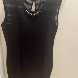 Svart kort klänning med pärldetalj - Snygg svart kort klänning med ärmlös design och transparenta ränder upptill. Rund halsringning med dekorativa pärlor och svarta stenar som ger extra bling. Perfekt för dig som gillar stilrena och eleganta plagg med en twist.