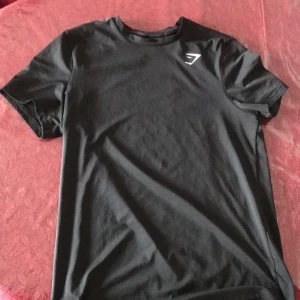 Svart Gymshark t-shirt S - Svart t-shirt från Gymshark i storlek S. Stilren design med Gymshark-logga i vitt på bröstet. Tillverkad i mjuk och stretchig polyesterblandning som sitter skönt mot kroppen. Perfekt för dig som gillar sportig och enkel stil.