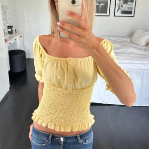 Gul offshoulder blus med knappar - Supersöt ljusgul blus med offshoulder och korta puffärmar. Smockad midja och volangkant nedtill ger en riktigt nice siluett. Framtill finns små knappar som detalj. Köpt på plick för dyrare pris🥰