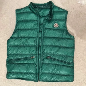 Grön dunväst från Moncler - Snygg grön dunväst från Moncler med quiltad design och klassisk logga på bröstet. Västen har två dragkedjeförsedda fickor framtill och hög krage. Perfekt att slänga över en hoodie för extra värme. Materialet är glansigt och känns riktigt lyxigt. Sjävklart äkta mvh Malte.202