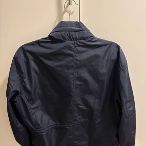 Mörkblå vindjacka från Bordelind - Snygg mörkblå vindjacka från Barchetta med hög krage och praktiska fickor framtill. Jackan har dragkedja och knappar, tillverkad i lätt polyester som passar perfekt när du vill ha en stilren look. Klassisk passform och långärmad design.