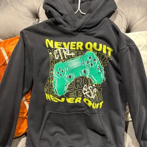 Svart hoodie med gaming-tryck H&M - Svart hoodie från H&M med stor grön och gul gamingkontroll på bröstet och texten 'Never Quit' i graffiti-stil. Tröjan har huva, magficka och långa ärmar. Perfekt för dig som älskar gaming och vill ha en chill streetstyle.