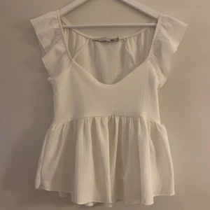 Vit blus med volangärmar - Supersöt vit blus med volangärmar och u- ringning. Toppen har en luftig, utsvängd passform nedtill och är tillverkad i ett lätt, strukturerat tyg. Perfekt att styla med jeans eller kjol för en fräsch look. Storlek S från Lager 157.