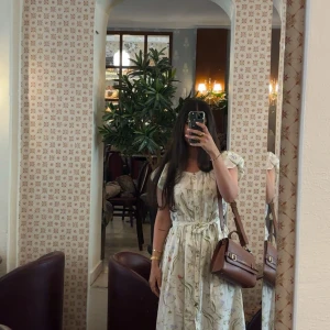 Blommig ljus långklänning med knyt - Säljer en superfin ljus långklänning med blommigt mönster och knyt i midjan. Klänningen har korta ärmar och en avslappnad passform som ger en romantisk vibe. Perfekt för varma dagar och har en mjuk, flowy känsla.