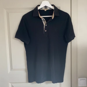 Svart Burberry piké - Svart t-shirt från Burberry med klassisk krage och knappslå med tre knappar. Kragen har Burberrys ikoniska rutiga mönster på insidan. T-shirten är i bomull och har korta ärmar samt en diskret broderad logga på bröstet.