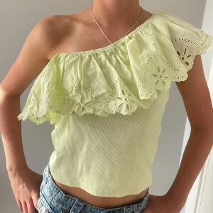 Limegrön one shoulder topp med volang - Superfin limegrön topp med one shoulder och bred volang med broderade detaljer. Toppen är ribbad i kroppen och har en luftig, blommig broderad volang som ger en romantisk vibe. Perfekt för dig som vill sticka ut med en unik och somrig look.