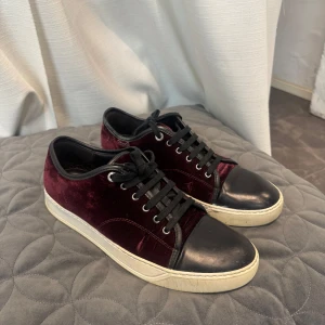 Vinröda sneakers från Lanvin i sammet - Snygga vinröda sneakers från Lanvin med ovandel i lyxig sammet och svart skinn på tån. Skorna har svarta snören, rund tå och vit platt sula. Perfekt för dig som vill sticka ut med en cool och exklusiv vibe.