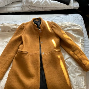 Senapsgul kappa med ståkrage - Säljer en snygg senapsgul kappa med ståkrage och knappar framtill. Jackan har två fickor och är helfodrad med svart tyg på insidan. Axelpartiet har en cool puffdetalj som ger extra stilpoäng. Perfekt för dig som vill sticka ut med färg i höst och vinter.