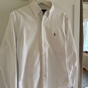 Vit skjorta från Ralph Lauren slim fit - Vit skjorta från Ralph Lauren i slim fit med klassisk krage och knappar hela vägen. Liten broderad logga på bröstet. Skjortan är långärmad och tillverkad i mjuk bomull. Perfekt för dig som gillar stilrena och tidlösa plagg.