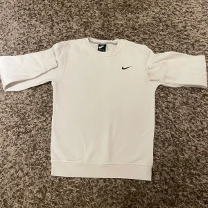 Vit sweatshirt från Nike - Säljer en stilren vit sweatshirt från Nike med klassisk svart logga på bröstet. Tröjan har rund hals och långa ärmar, perfekt för chill dagar eller när du vill ha en clean look. Materialet är mjukt och skönt, passar dig som gillar enkel och sportig stil.