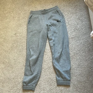 Grå mjukisbyxor från Puma - Snygga grå mjukisbyxor från Puma med klassisk svart logga på benet. Byxorna har elastisk midja, ribbade muddar vid benslut och två sidofickor. 