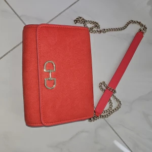 Röd axelväska med kedja - Snygg röd axelväska med guldfärgad kedja och metallspänne framtill. Väskan har en stilren, rektangulär form och är gjord i mocka med detaljer i skinn. Perfekt för att lyfta din outfit med en pop av färg.