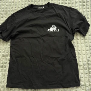 Svart Amiri t-shirt med ängelprint - Svart t-shirt från Amiri i mjuk bomull. Framsidan har vit Amiri-logga och en liten ängel, medan baksidan har ett stort vitt print med en detaljerad ängel och loggan. Klassisk rund hals och korta ärmar. Perfekt för dig som gillar streetwear med unik design.
