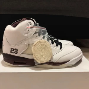 Nike Jordan 5 A Ma Maniére Burgundy Violet ore  - Nike Air Jordan 5 A Ma Maniére sneakers i vit och vinröd med svart snörning och extra vita skosnören. Skorna har högt skaft, broderad 23 på sidan och Nike Air-logga bak. Materialet är skinn med meshpaneler och detaljer i vinrött på sula och plös.