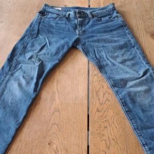 Levi's 502 blå jeans W29 L32 - Snygga blå Levi's 502 jeans i storlek: W29/L32 med en klassisk femficksdesign och rakt snitt. Jeansen har en brun läderpatch bak och den ikoniska röda Levi's-taggen vid bakfickan. Tillverkade i slitstarkt bomullsjeansmaterial, perfekta för en chill och stilren look. Jeansen är i väldigt bra skick, de är skrynkliga pga att de har legat i garderoben ett tag utan användning.