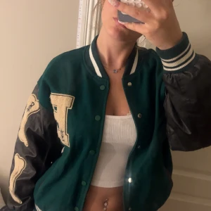 Grön och svart bomberjacka med broderier - Säljer en grön och svart bomberjacka med stora broderade bokstäver och ben på ärmarna. Jackan har ribbade muddar med vita ränder, tryckknappar och svarta detaljer vid fickorna. Perfekt för dig som vill sticka ut med en cool streetstil! 😇